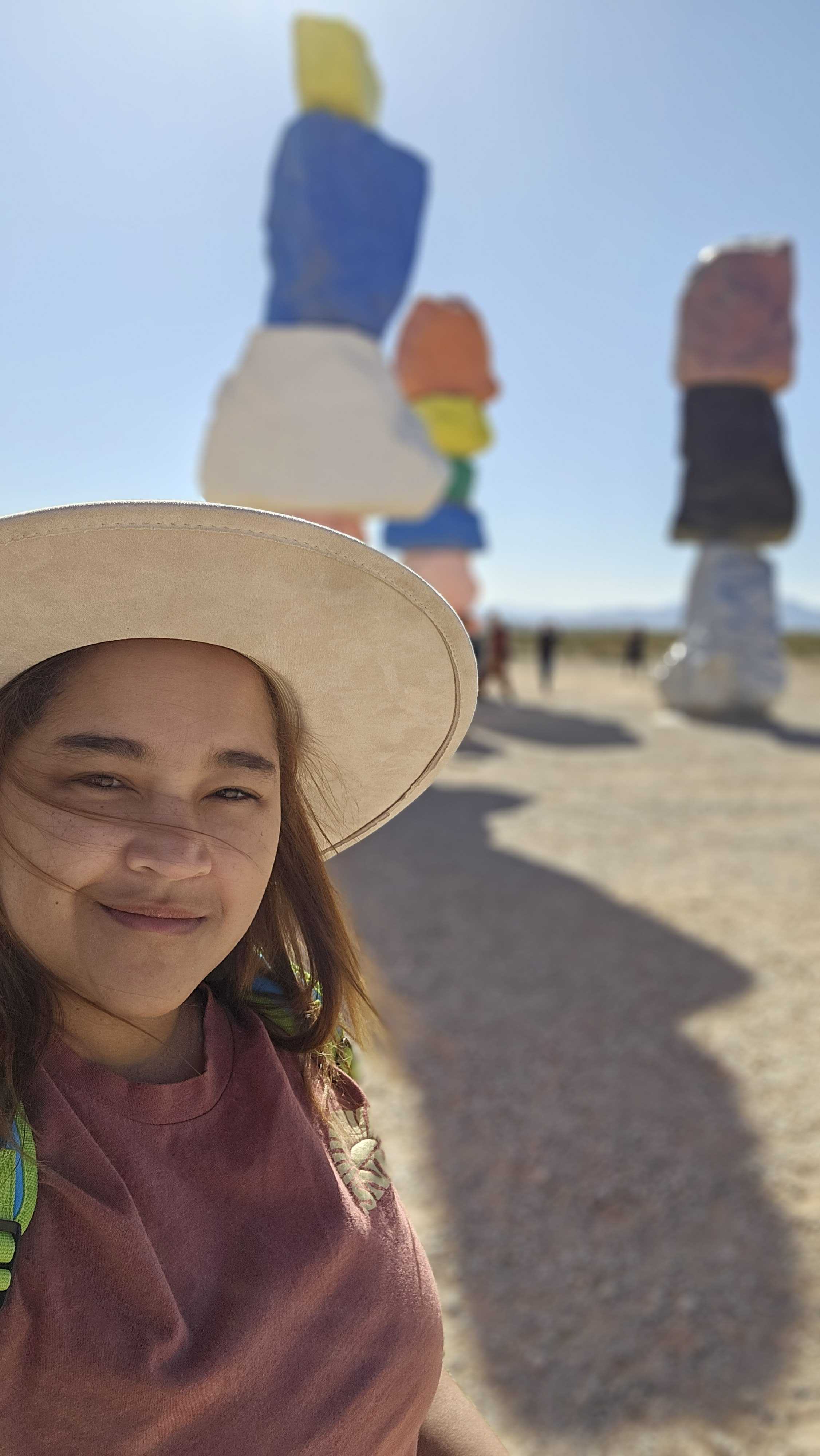A woman in a beige, wide‑brimmed hat smiles softly at the camera in a sunlit desert. Behind her, the brightly colored stacked boulders of the Seven Magic Mountains art installation rise slightly out of focus.